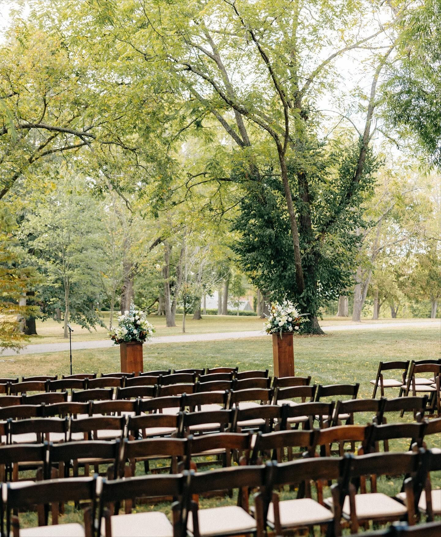 Outdoor wedding ceremony setup with rows of brown chairs facing floral arrangements and trees.