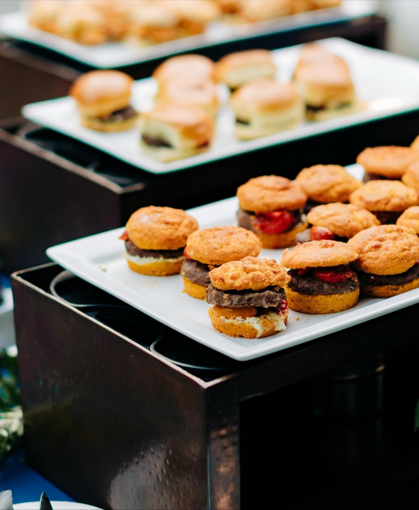 Mini burgers on white platters, stacked on dark brown boxes, set out for a gathering.