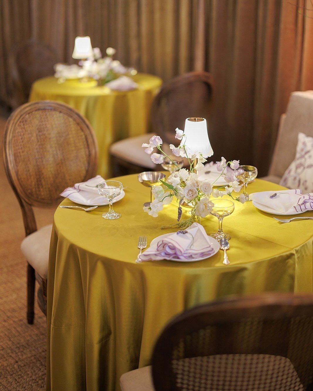 Round tables with yellow tablecloths, white flowers, lamps, and place settings. Wooden chairs surround the tables.