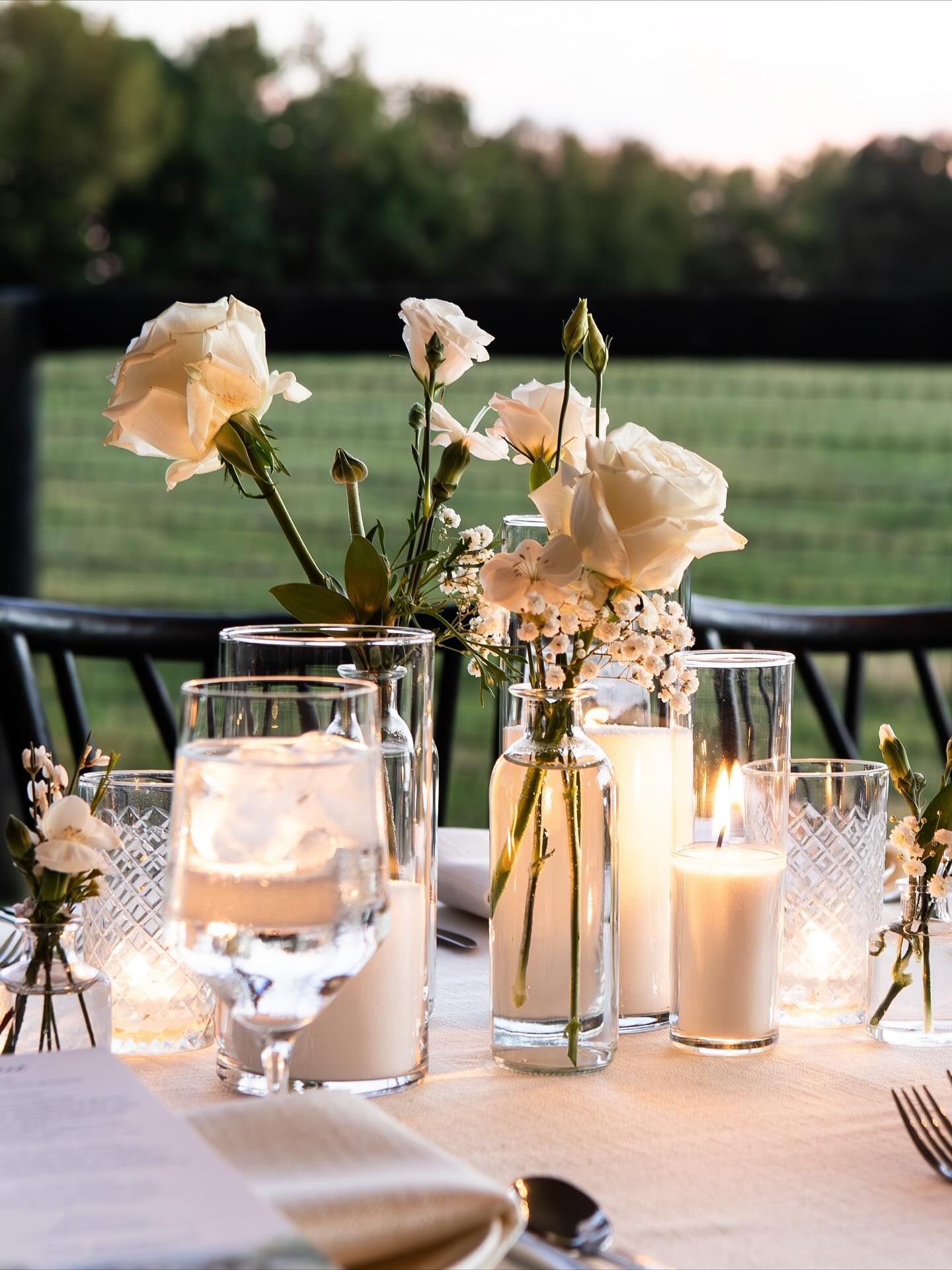 Table setting with white flowers, candles, and water glasses outdoors, with a blurred natural backdrop.