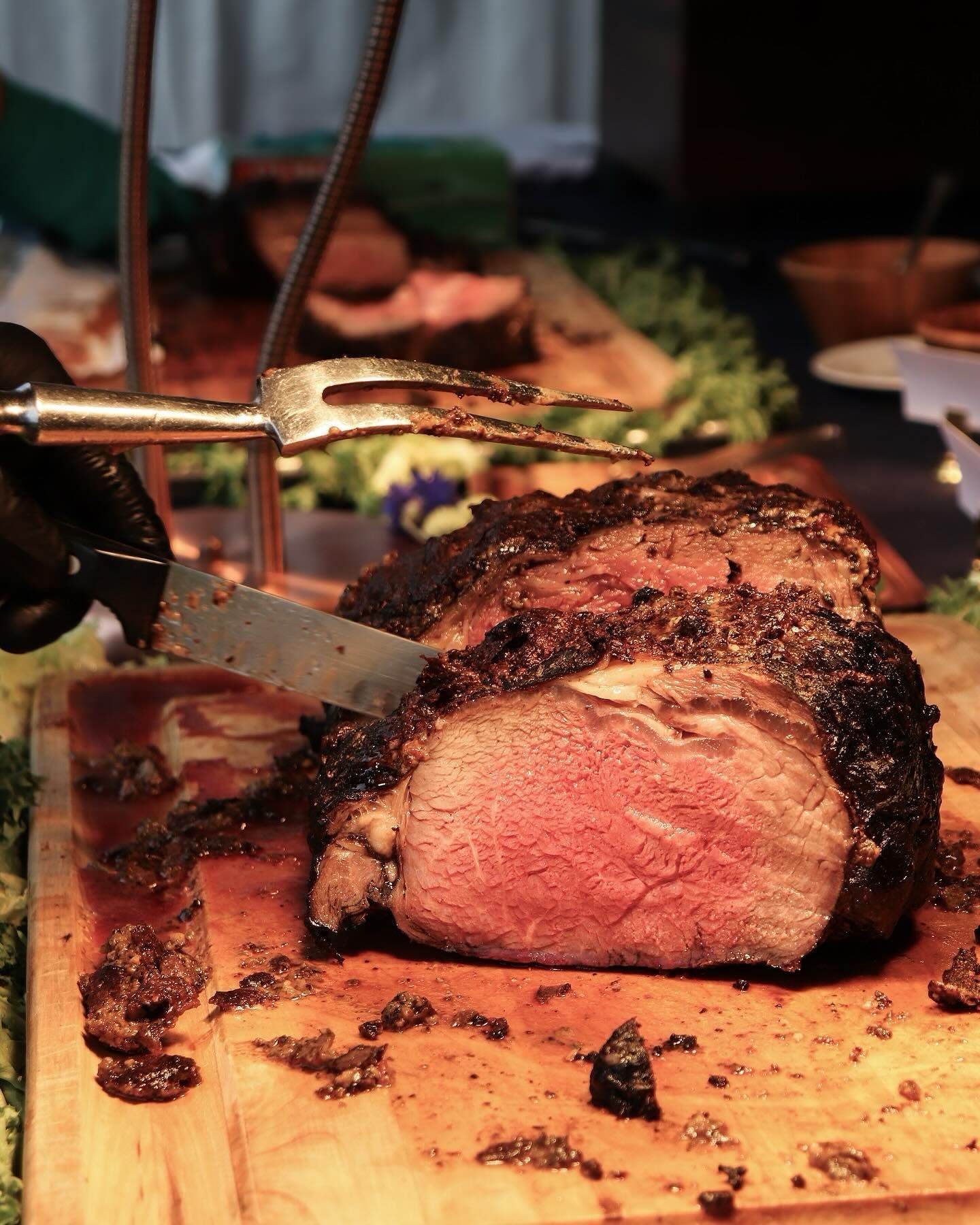A gloved hand uses a knife and fork to slice a medium-rare roast on a wooden cutting board.