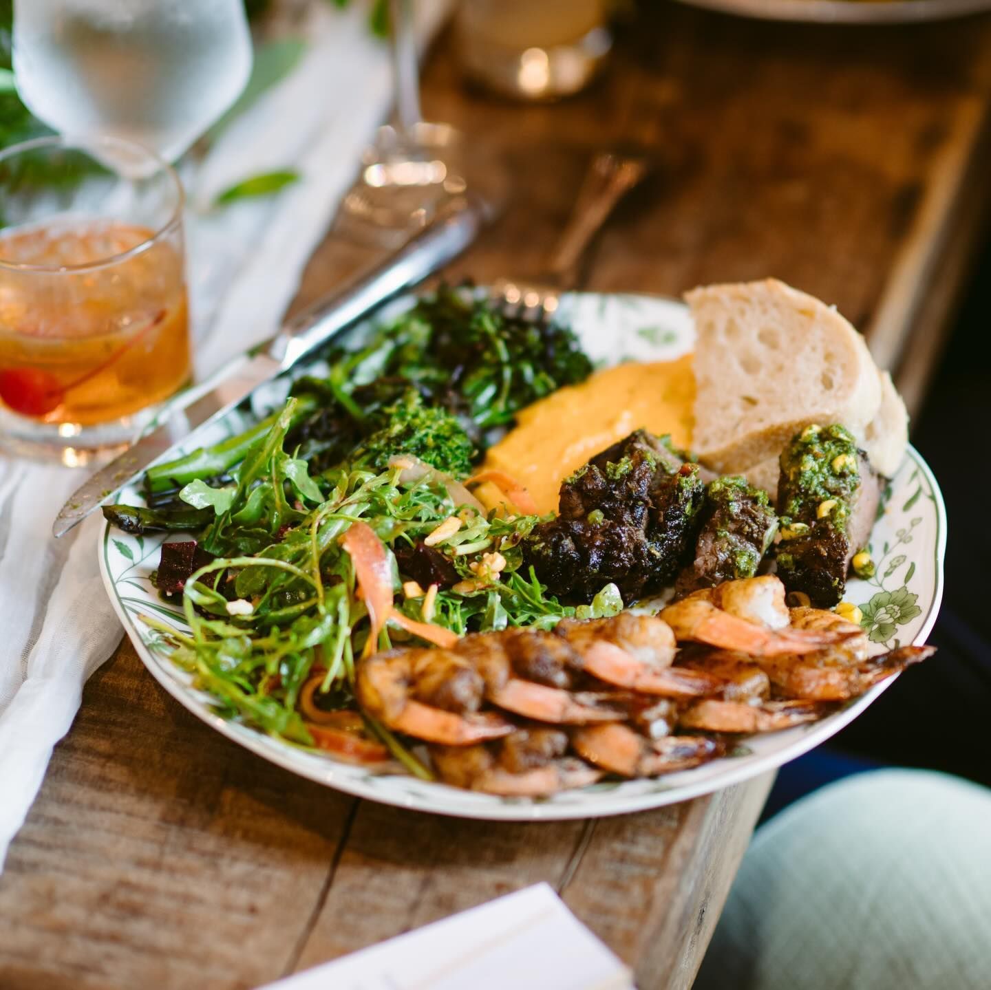 A plated meal of shrimp, salad, dark roasted vegetables, a dollop of yellow purée, and bread on a rustic wooden table.