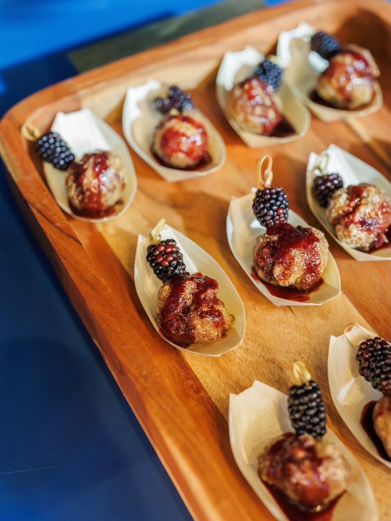 A wooden serving tray holds several individual leaf-shaped boats, each containing a meatball topped with a blackberry.
