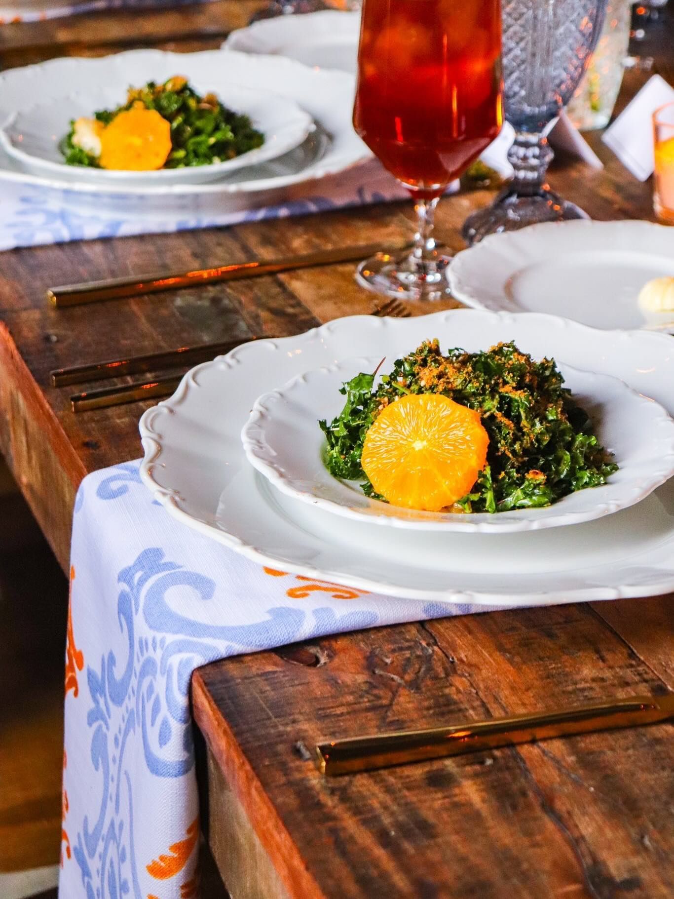 A rustic wooden table is set with white scalloped plates featuring kale salads topped with orange slices and red drinks.