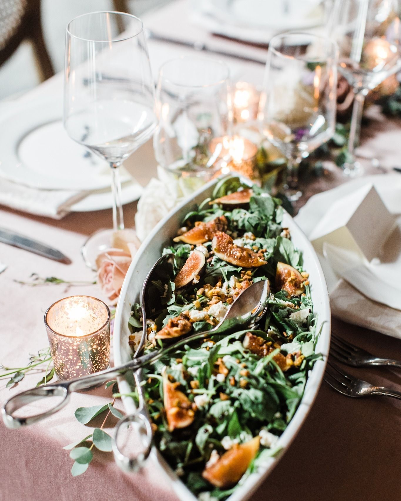 A salad with figs and greens in an oval serving dish on a wedding table with wine glasses and glowing votive candles.