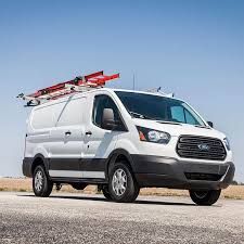 White Ford Transit van with ladder rack, parked on pavement, blue sky.