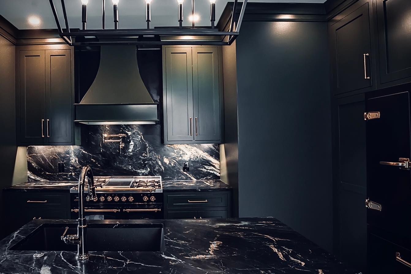 Dark kitchen with black cabinets, marble countertops, stainless steel appliances, and a modern light fixture.