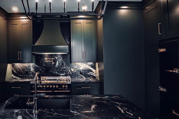 Dark kitchen with black cabinets, marble countertops, stainless steel appliances, and a modern light fixture.