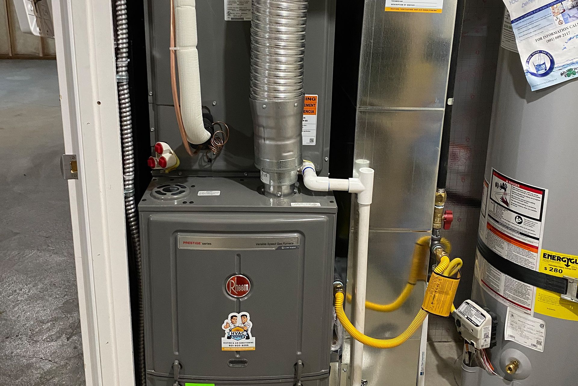 Furnace and water heater in a utility room. Gray furnace, silver ductwork, and white pipes are visible.