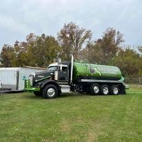 A green tanker truck is parked in a grassy field next to a trailer.