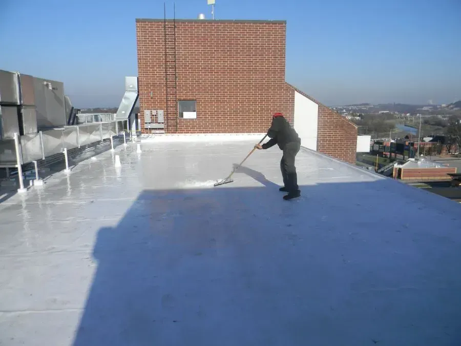 A person spreads a white liquid coating across a commercial rooftop using a long-handled brush on a sunny day.