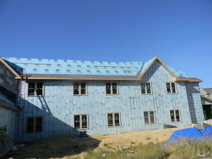 A two-story building under construction, covered in blue protective sheathing and underlayment with installed windows.
