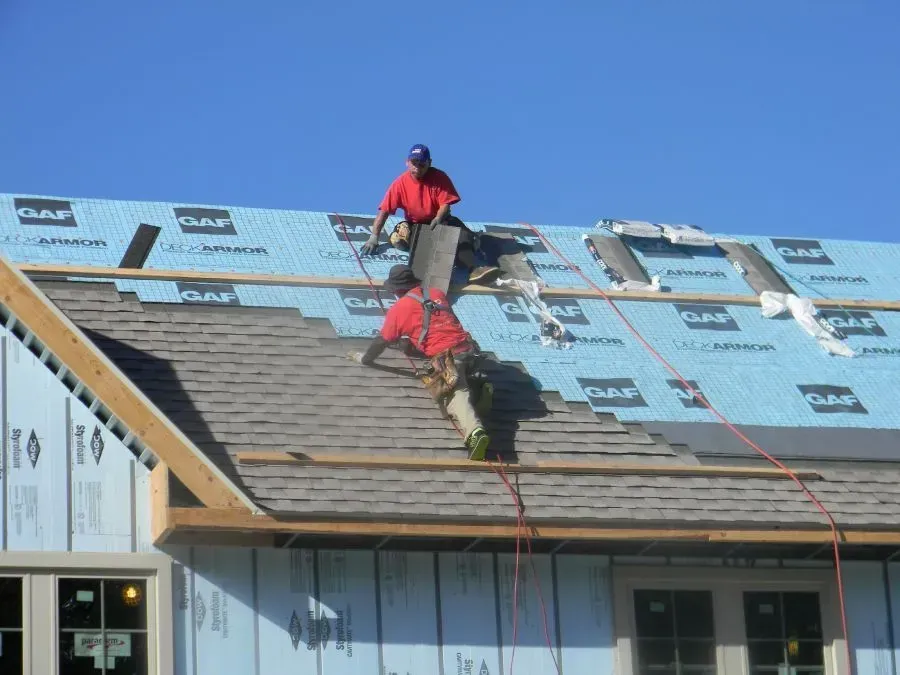 Two workers in red shirts install shingles on a residential roof covered with GAF brand blue underlayment.