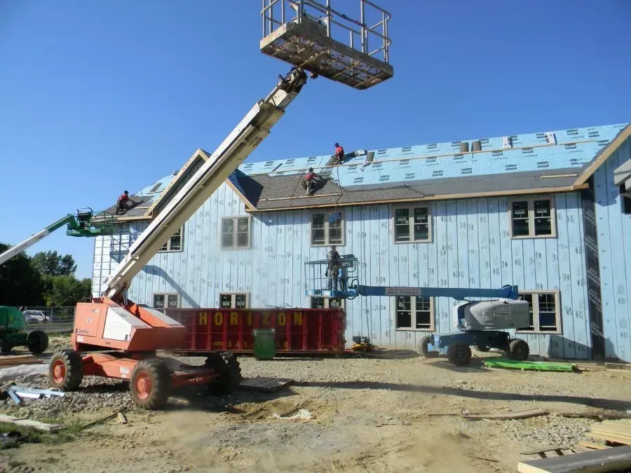 Workers install roofing on a building under construction, assisted by two orange and blue boom lifts in a dirt lot.