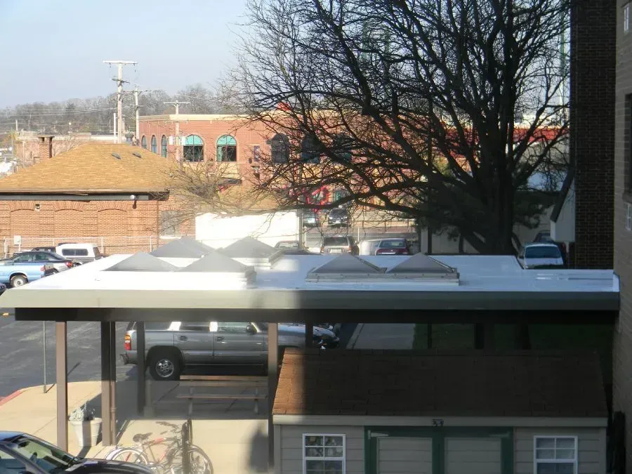 A white rooftop with skylights over a parking area, with a storage shed in the foreground and brick buildings in the back.