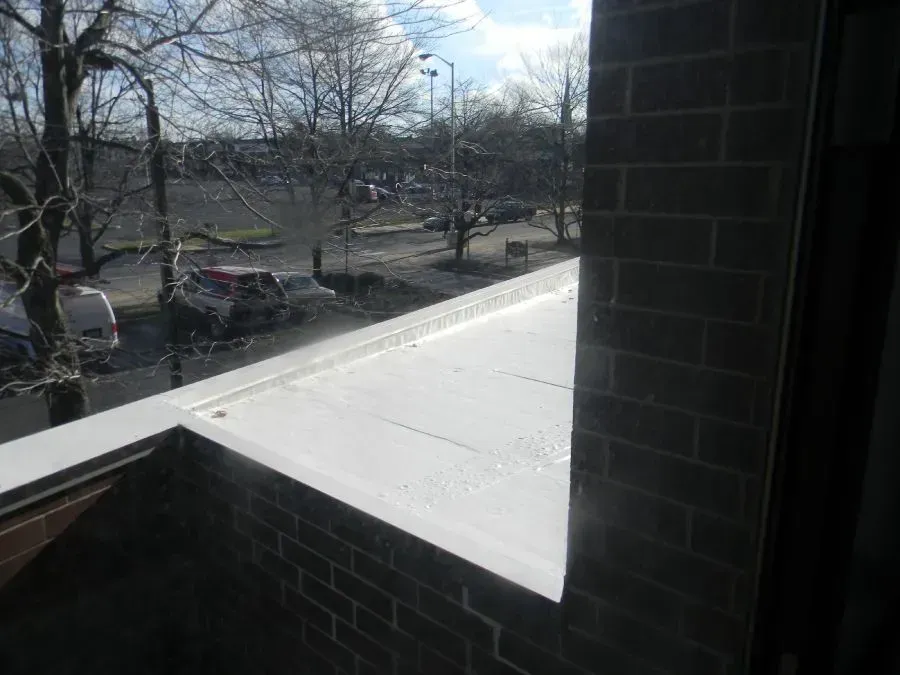 A white flat roof surface seen from a window, overlooking a parking lot with trees and cars on a sunny day.