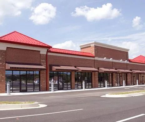 A modern, empty red-brick retail strip mall with a red metal roof and several glass storefronts under a blue sky.