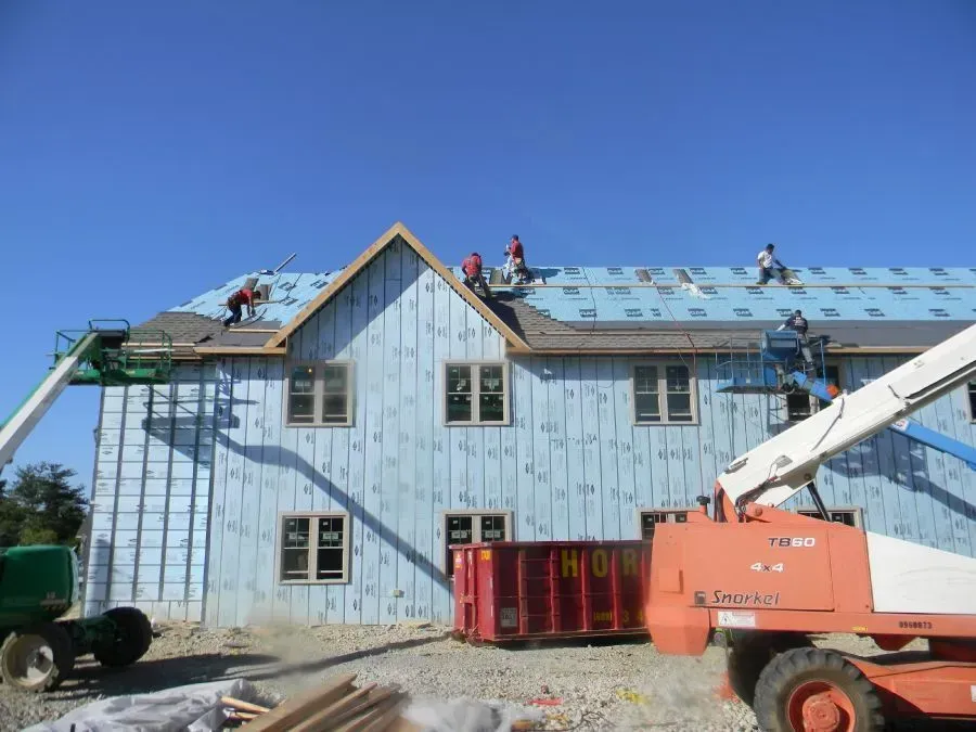 Construction workers install roofing on a two-story building covered in light blue exterior insulation.