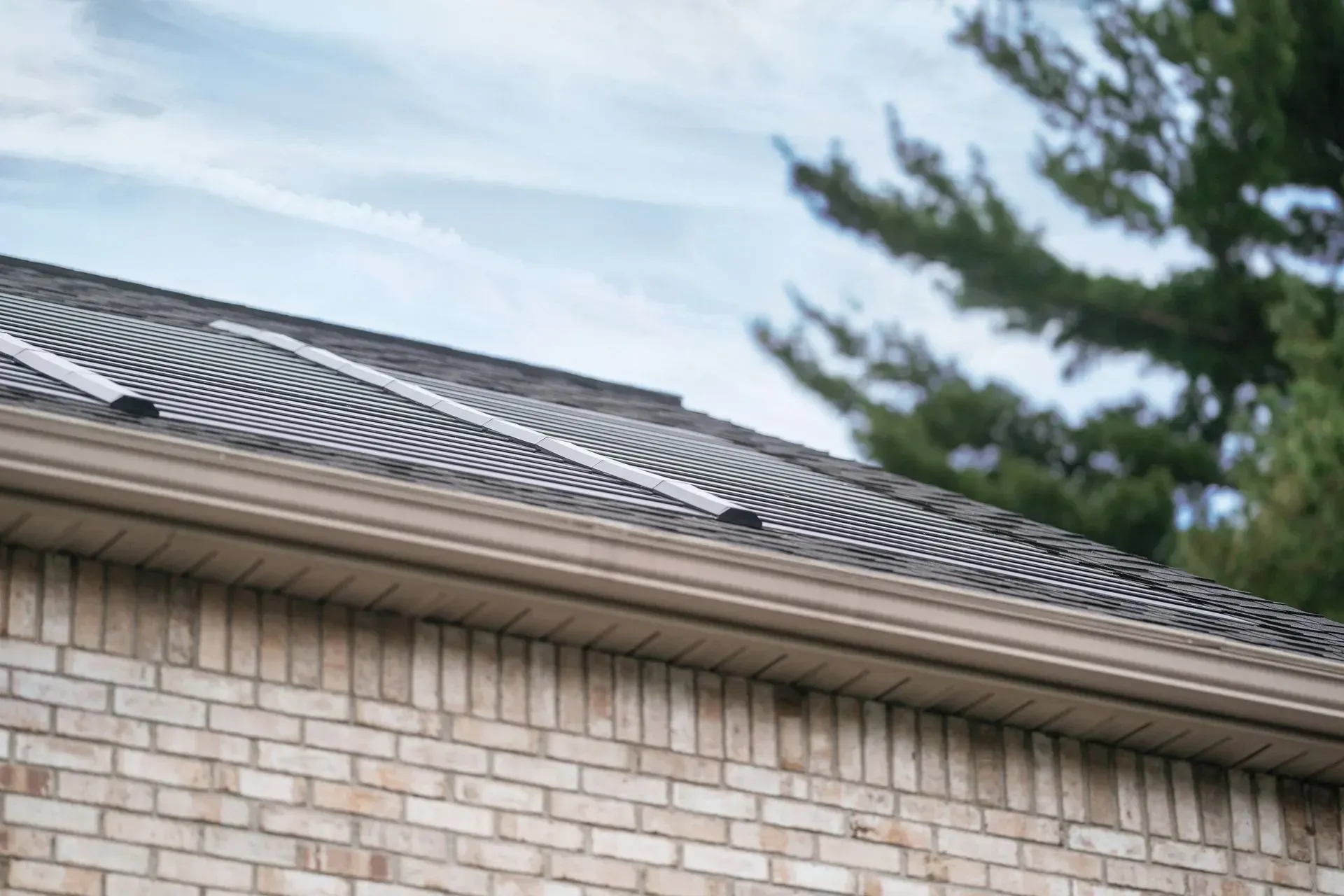 A low-angle view of a brick house roof with solar heating mats installed over shingles, next to green pine trees.