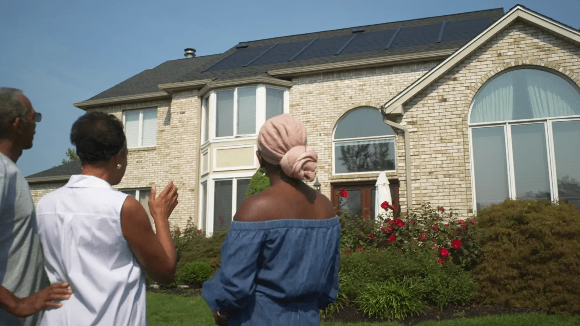 Three people stand in a yard looking at a two-story stone house with solar panels on the roof.