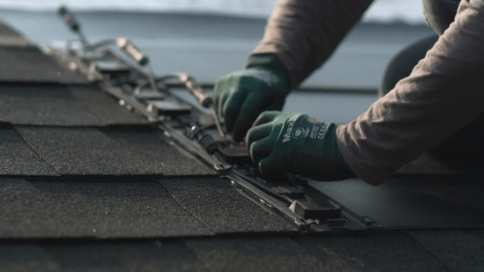 A person in green work gloves installs metal mounting hardware on a dark, shingled roof.