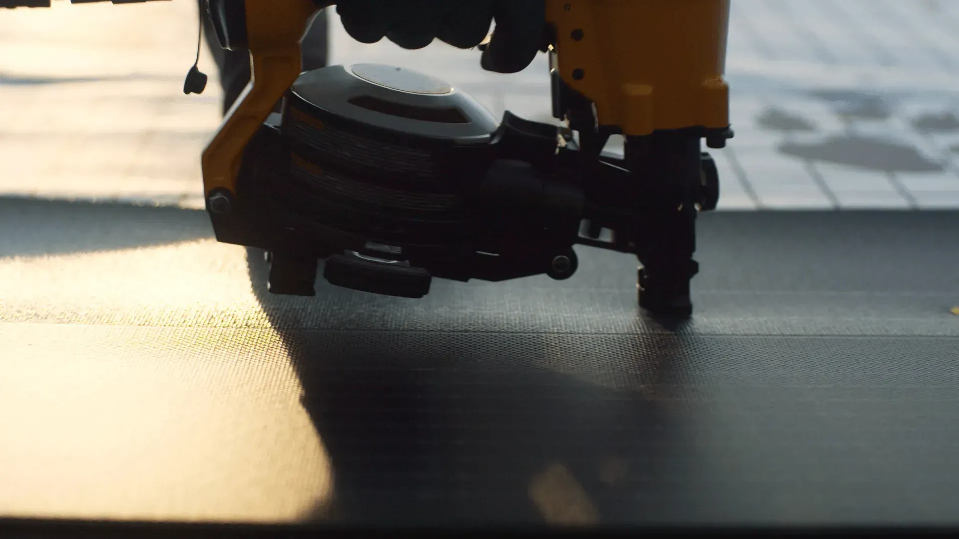 A yellow pneumatic nail gun pressing into a black roofing material under sunlight.