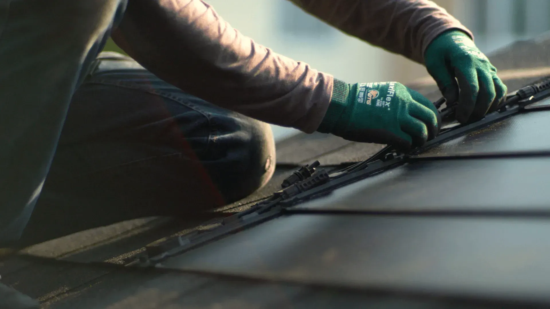A person wearing green work gloves installs dark, rectangular solar roof tiles.