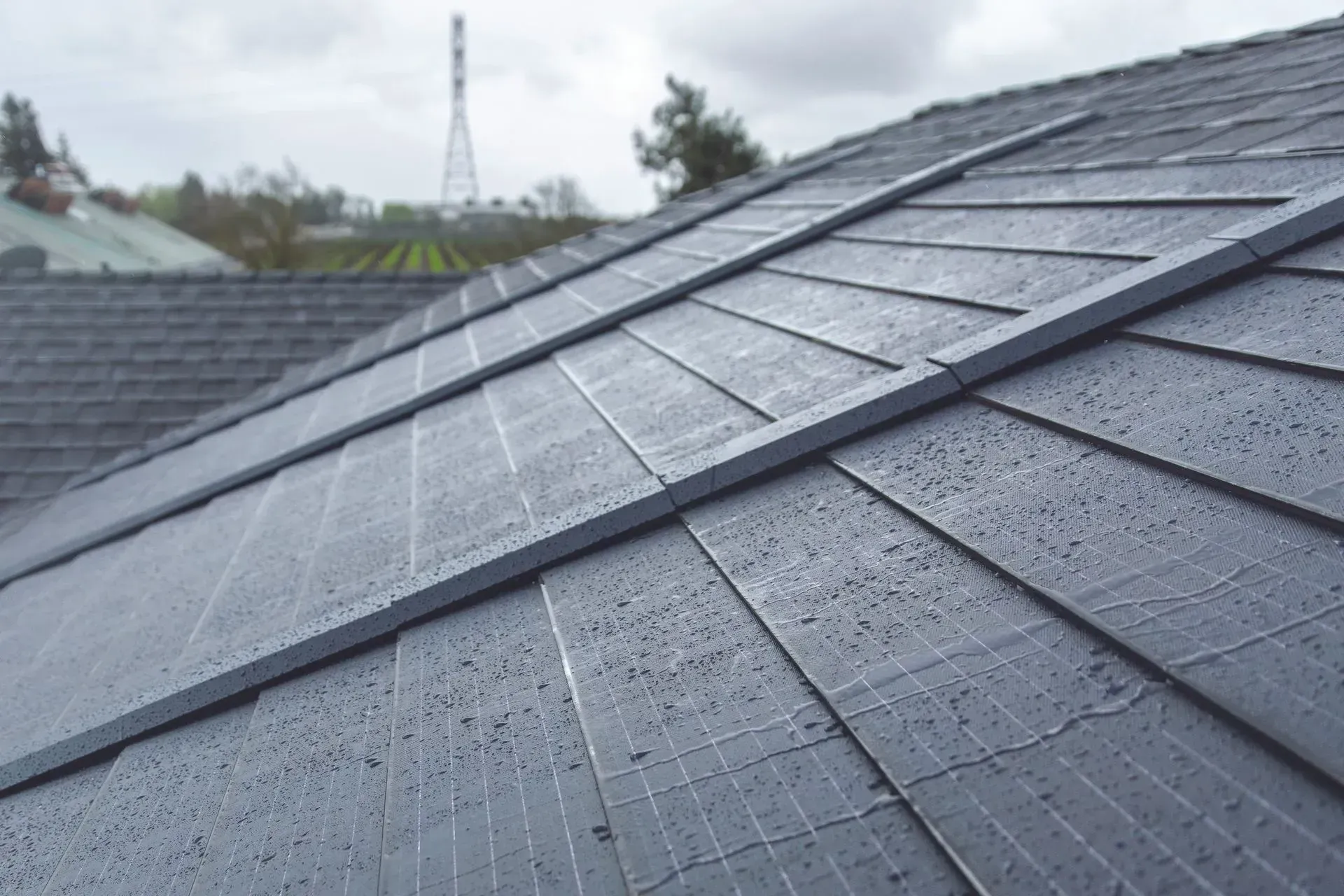 Solar roof shingles covering a slanted roof with a distant utility tower in the background.