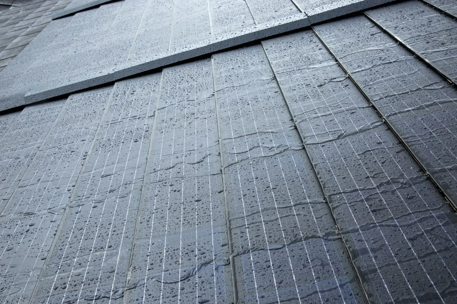 Solar shingles on a roof with visible rain droplets on their textured, grey-black surfaces.
