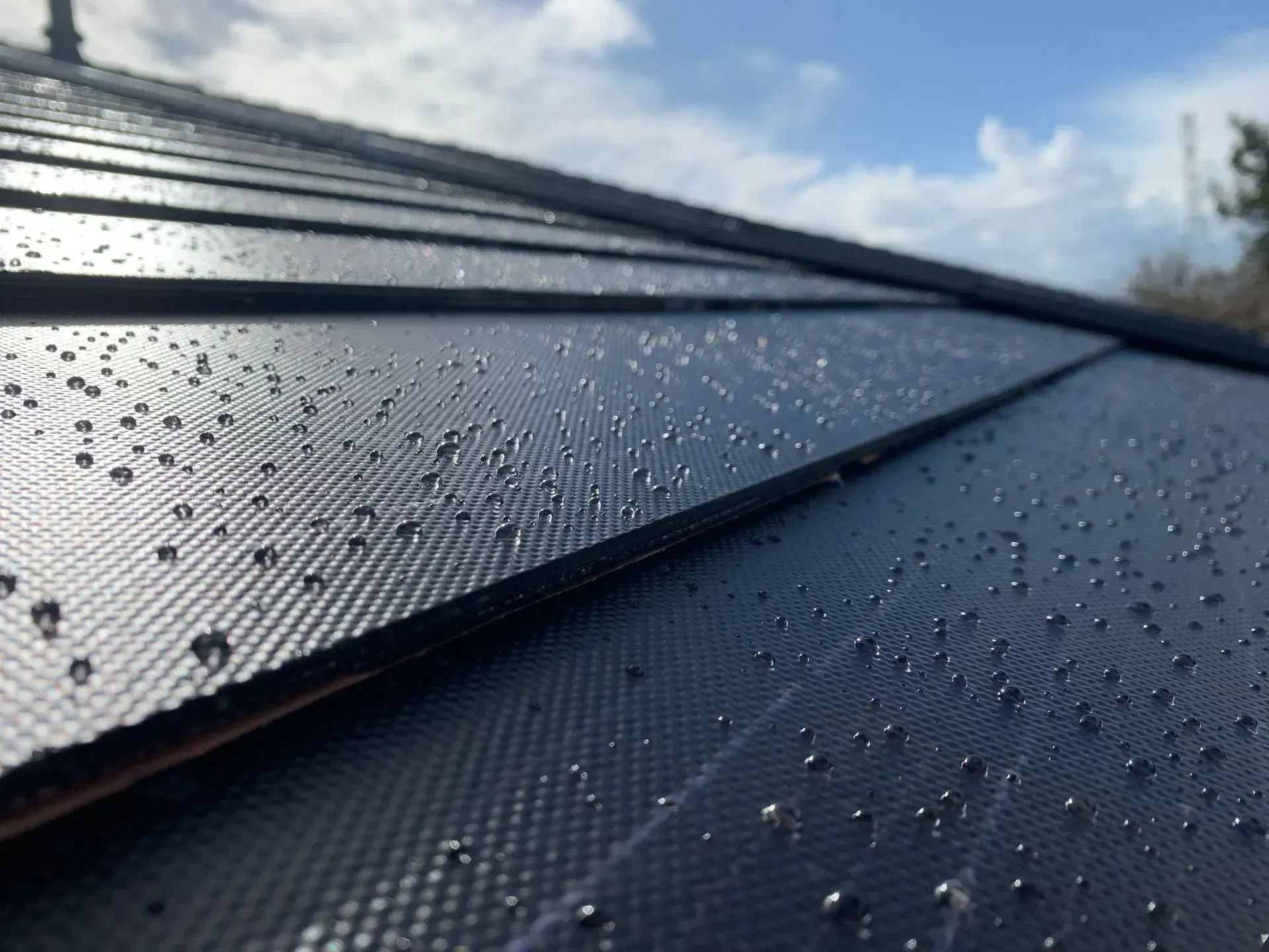 Close-up of raindrops resting on the textured, dark, sloped surface of solar roof tiles under a blue, cloudy sky.