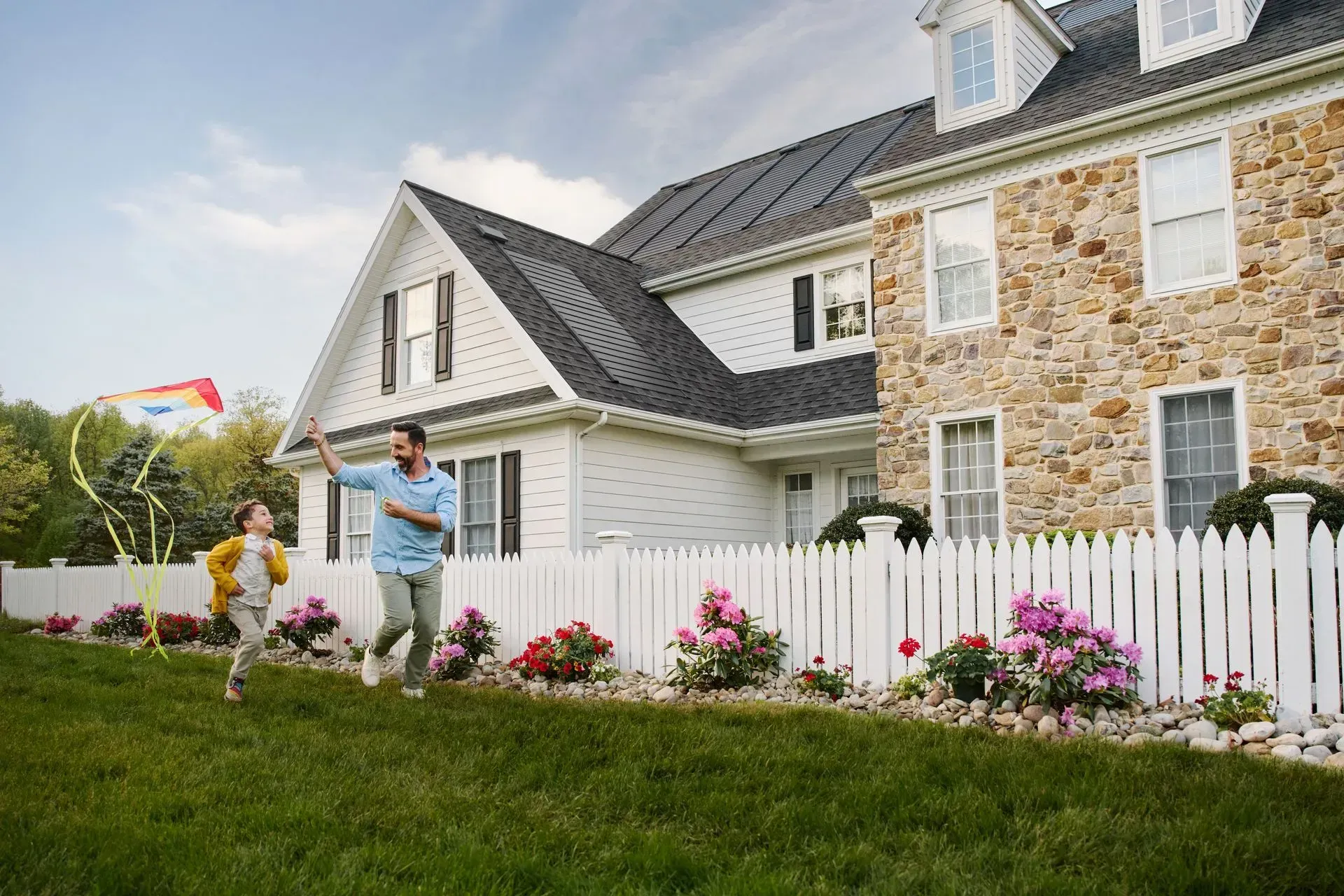 A parent and child run across a grassy lawn, flying a colorful kite in front of a white house with a stone facade.