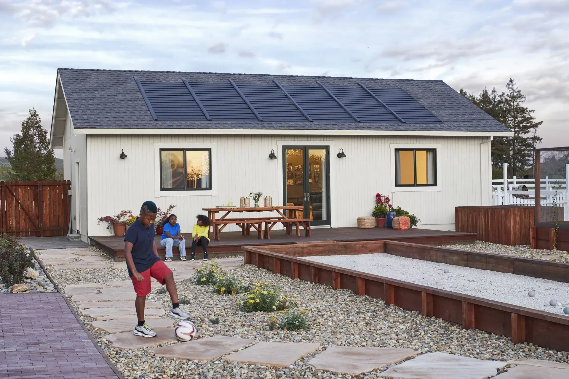 A white house with solar panels on the roof sits behind a gravel yard with a bocce court and people relaxing on a deck.