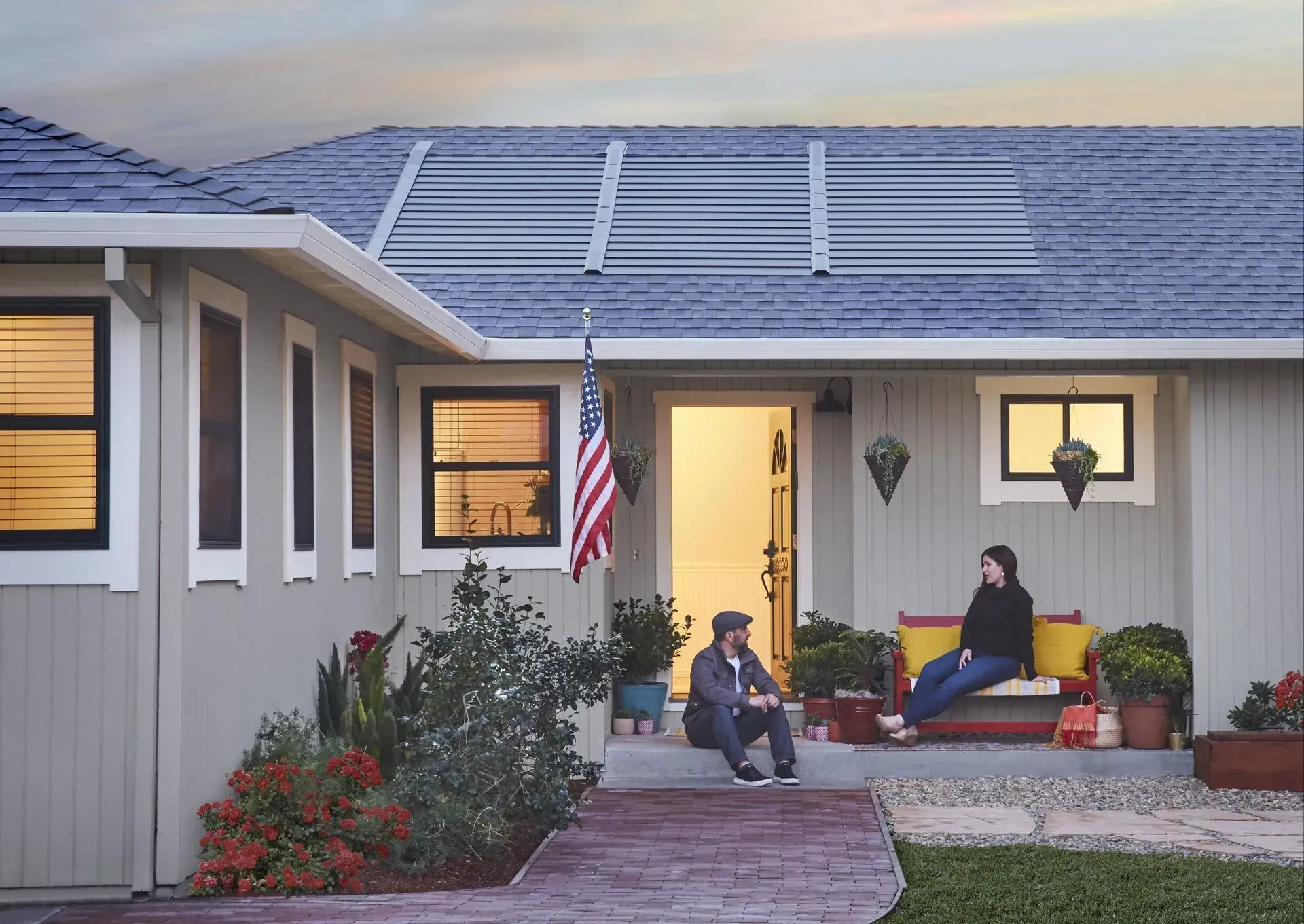 Two people sit on the porch of a grey house with solar panels on the roof and an American flag near the front door.