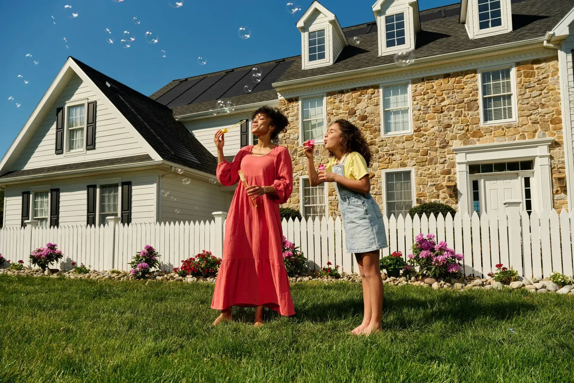 A woman in a red dress and a girl in overalls blow bubbles in front of a stone house with a white picket fence.