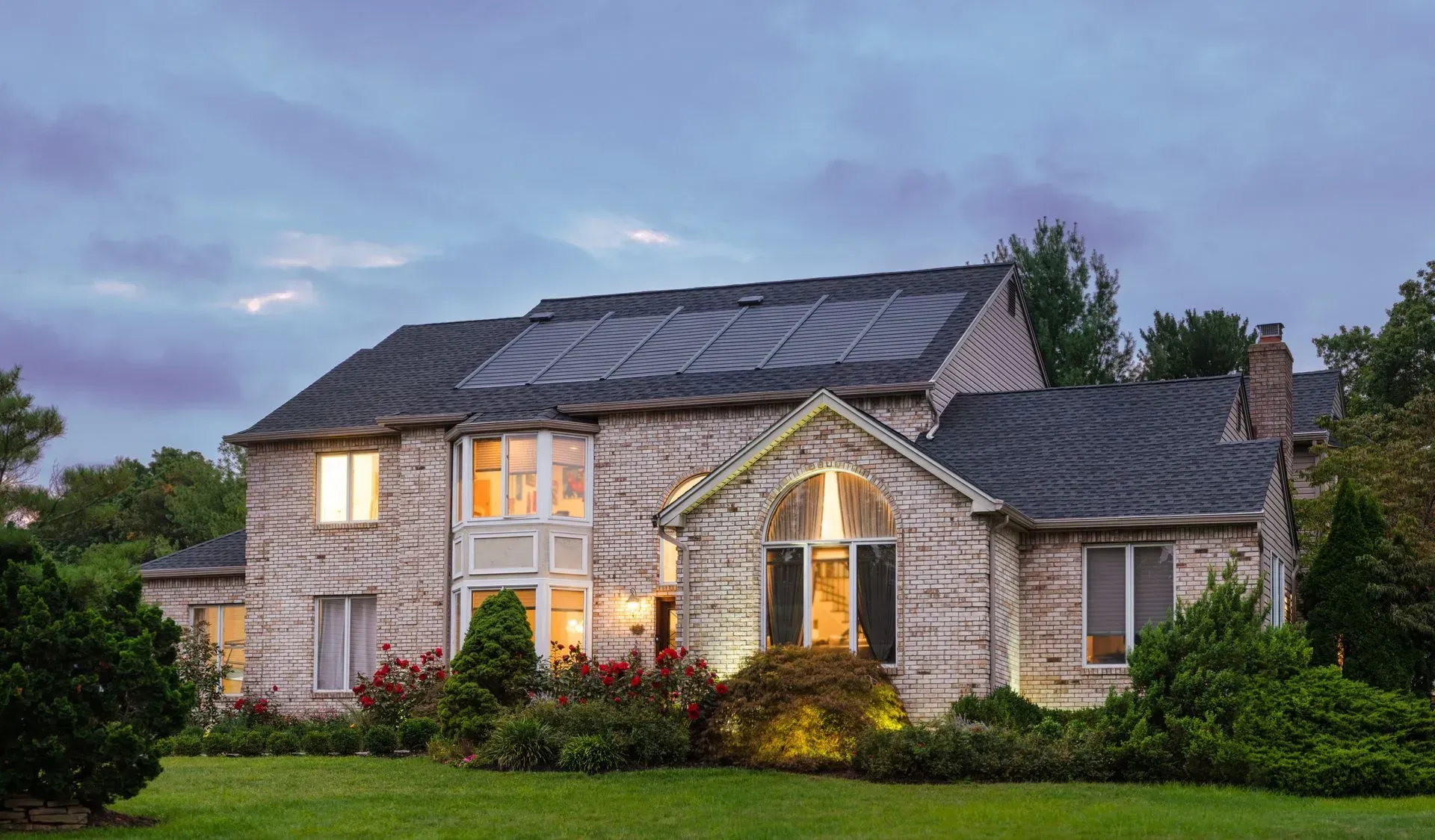 A stone, two-story suburban house with a dark shingled roof featuring solar panels, surrounded by trees and landscaping.