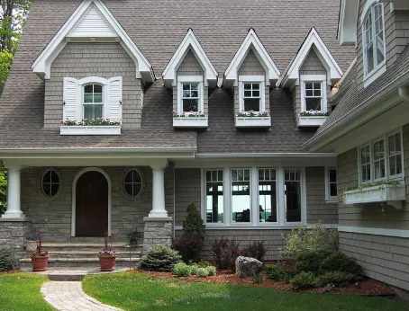 A tan, multi-story house with shingle siding, a front porch, and four dormer windows, featuring a stone entry and lawn.