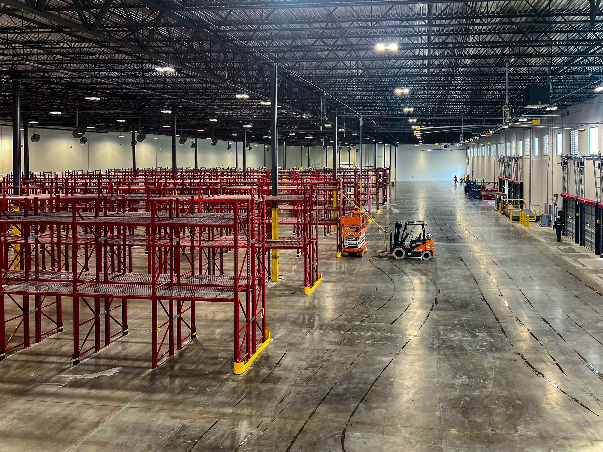 Empty warehouse with red shelving, forklifts, and workers. Black ceiling, concrete floor.