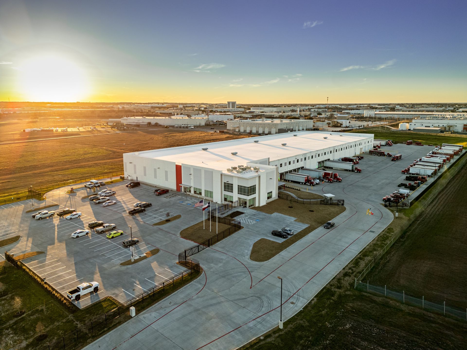 Warehouse at sunset with trucks, cars, and surrounding industrial area.