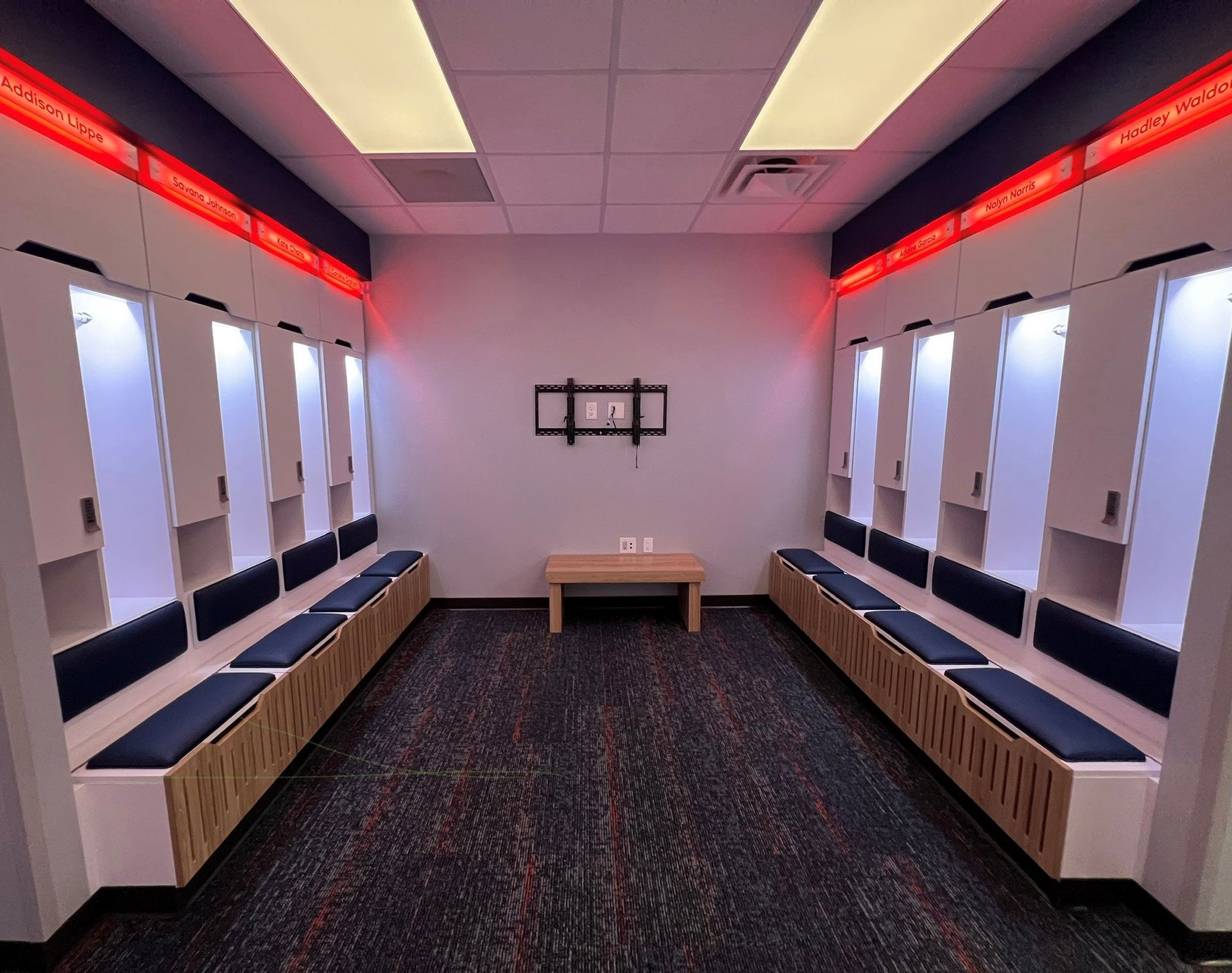 Locker room with white lockers, blue benches, a small table, and a mounted TV. Red and white lighting.