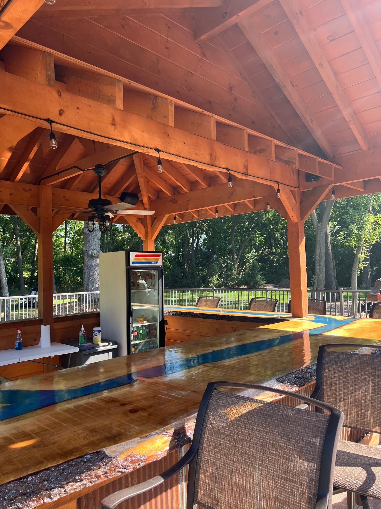 Covered outdoor pavilion with wooden beams, picnic tables, and a wet floor beside a park's grassy trees.