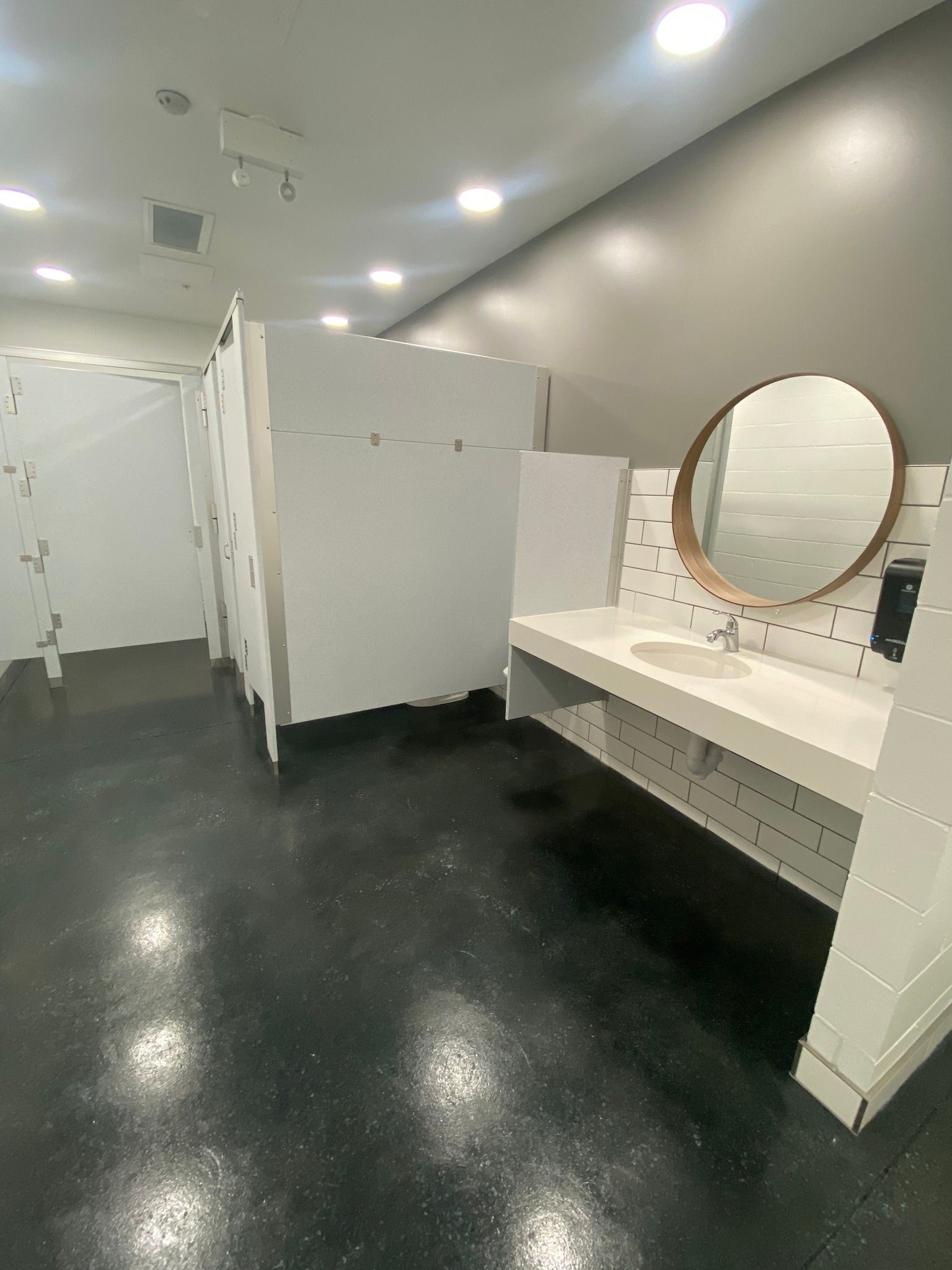 Modern restroom interior with white stalls, black floor, and a vanity with round mirror.