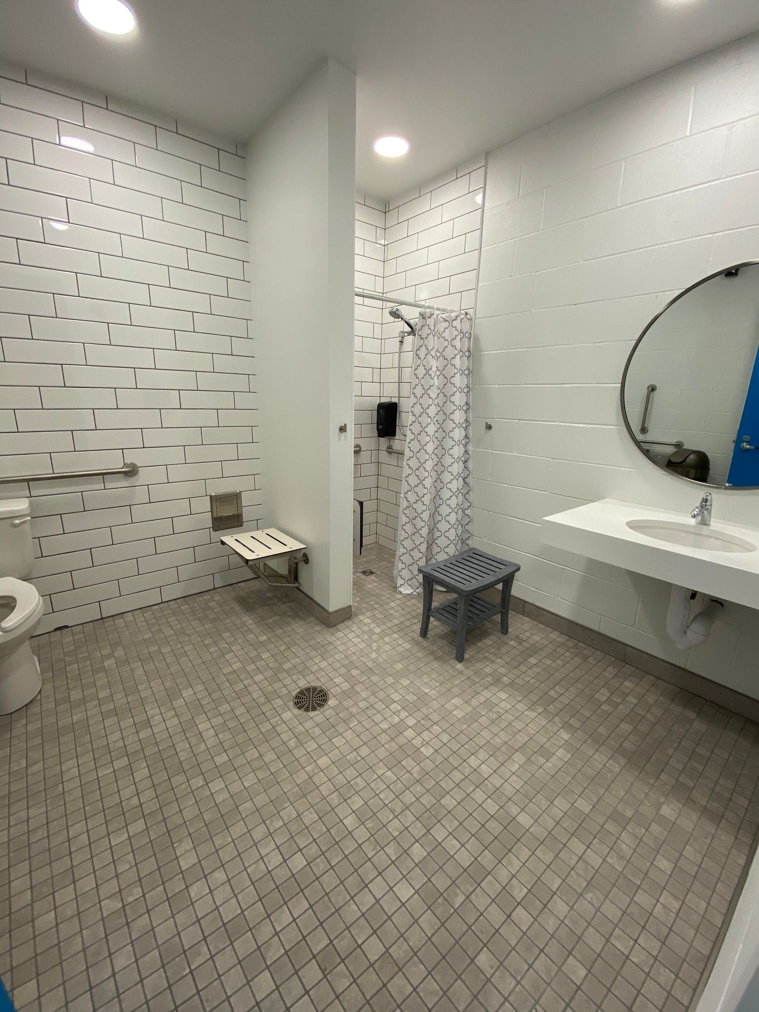 Accessible restroom with white tiled walls, patterned floor, sink, mirror, toilet, and shower area