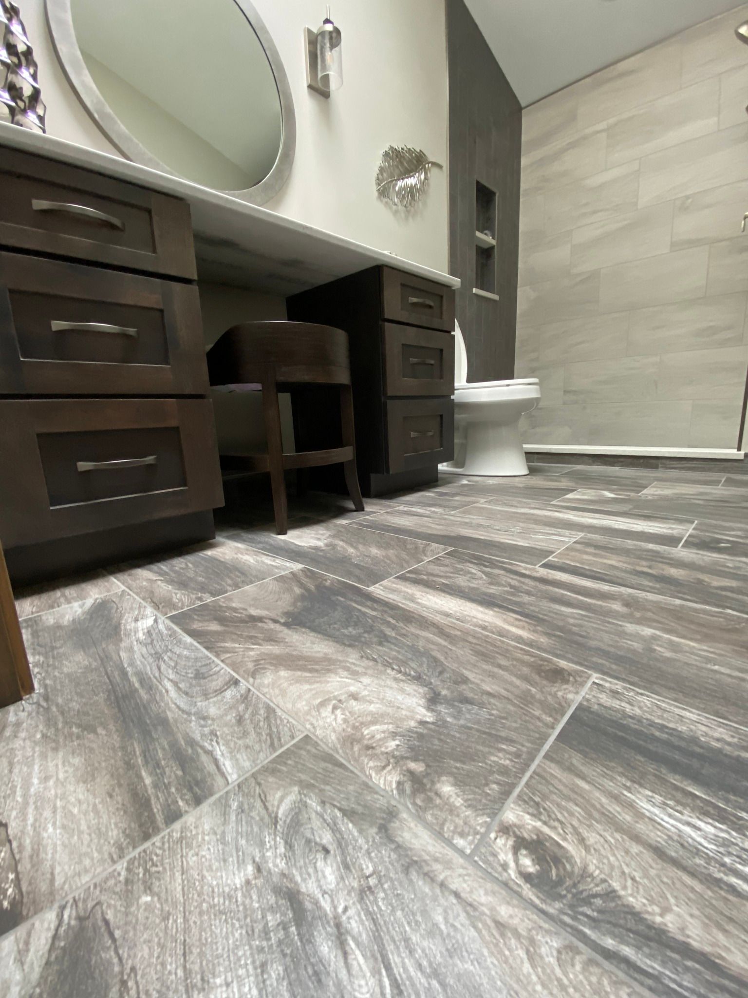 Modern bathroom with dark wood vanity, round mirror, tiled shower, and gray wood-look flooring.