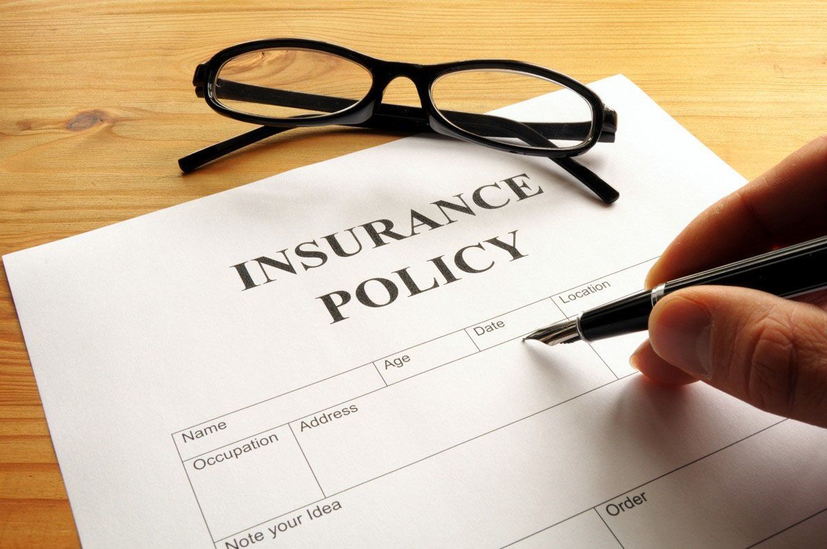 A hand holds a pen over an insurance policy form, with eyeglasses resting on the document on a wooden desk.