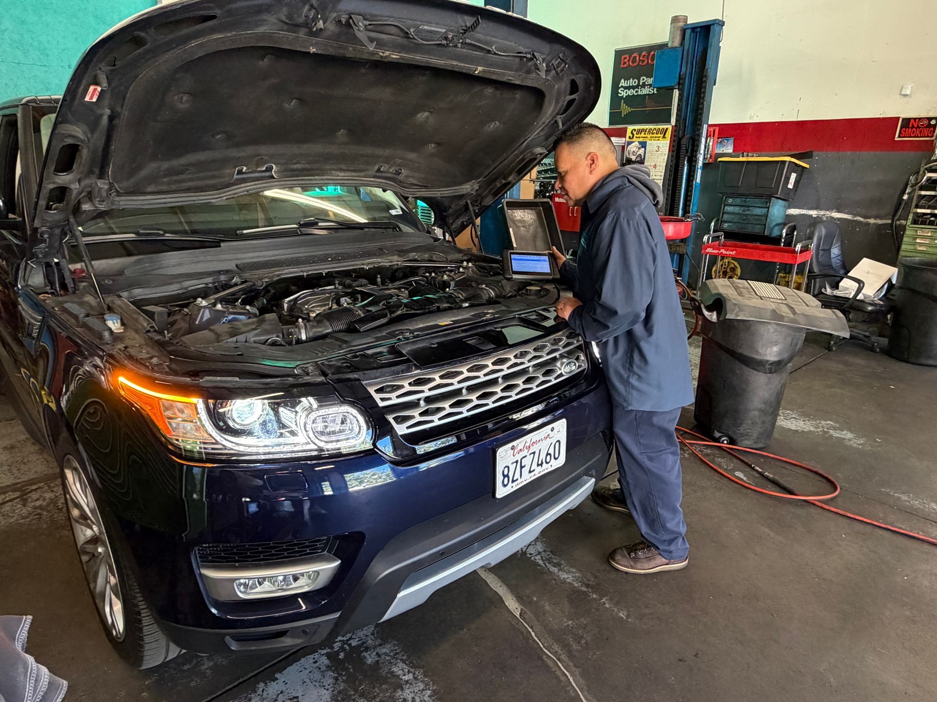 Mechanic in blue jumpsuit checks a blue Range Rover with open hood in a garage, using a laptop.