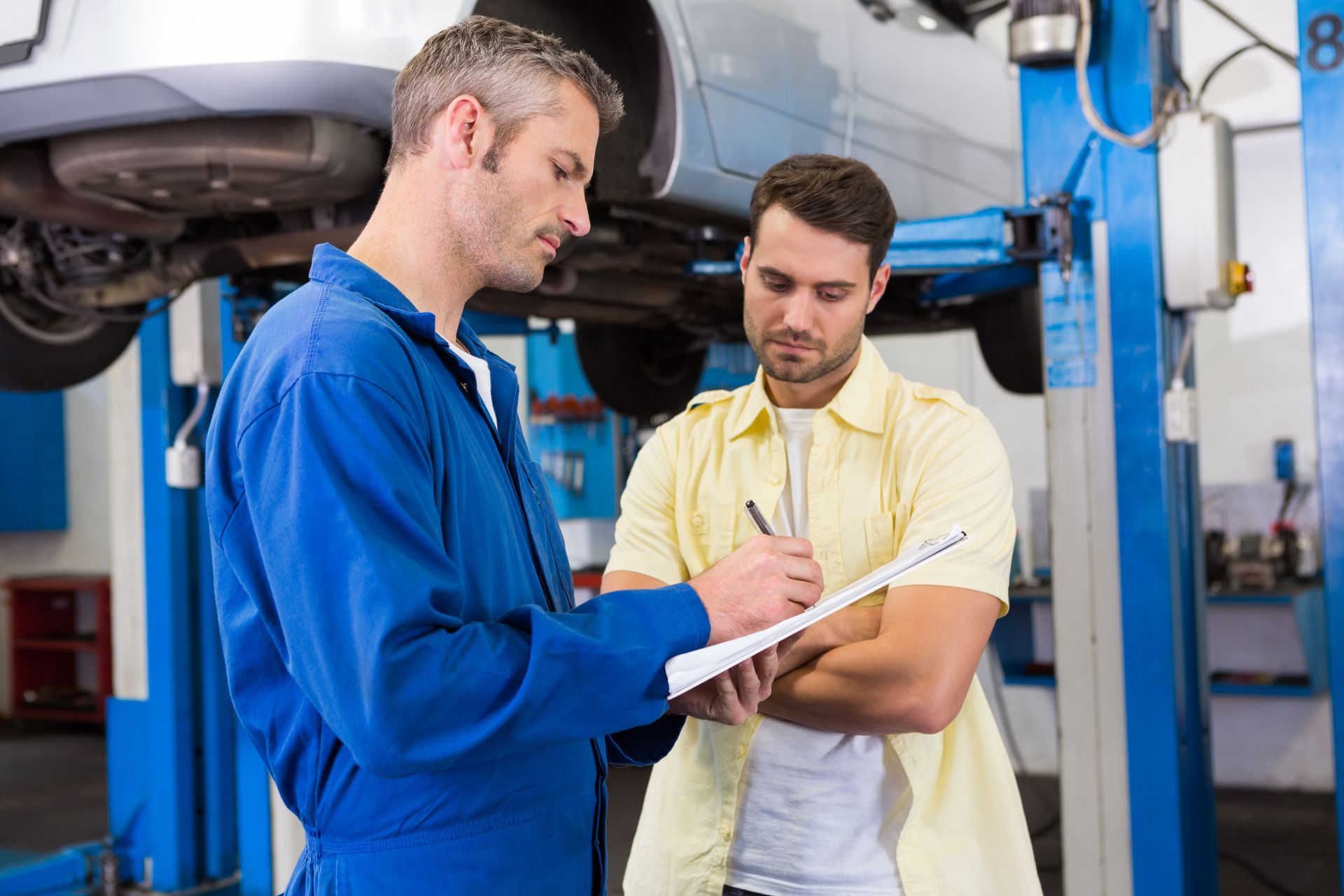 Mechanic in blue jumpsuit shows clipboard to customer in yellow shirt; car on lift in shop.