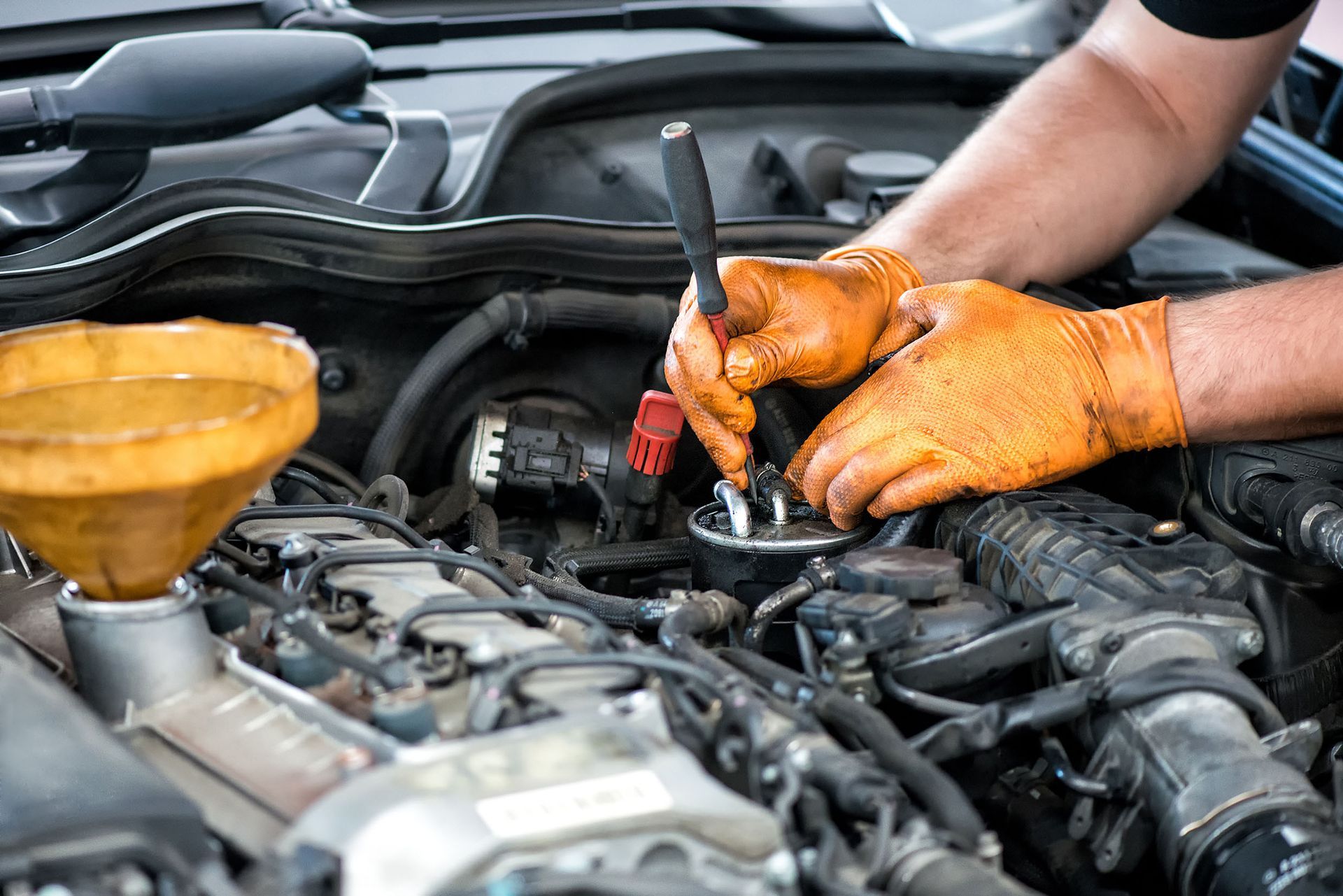Mechanic in orange gloves working on car engine, holding tool near components with a funnel visible.