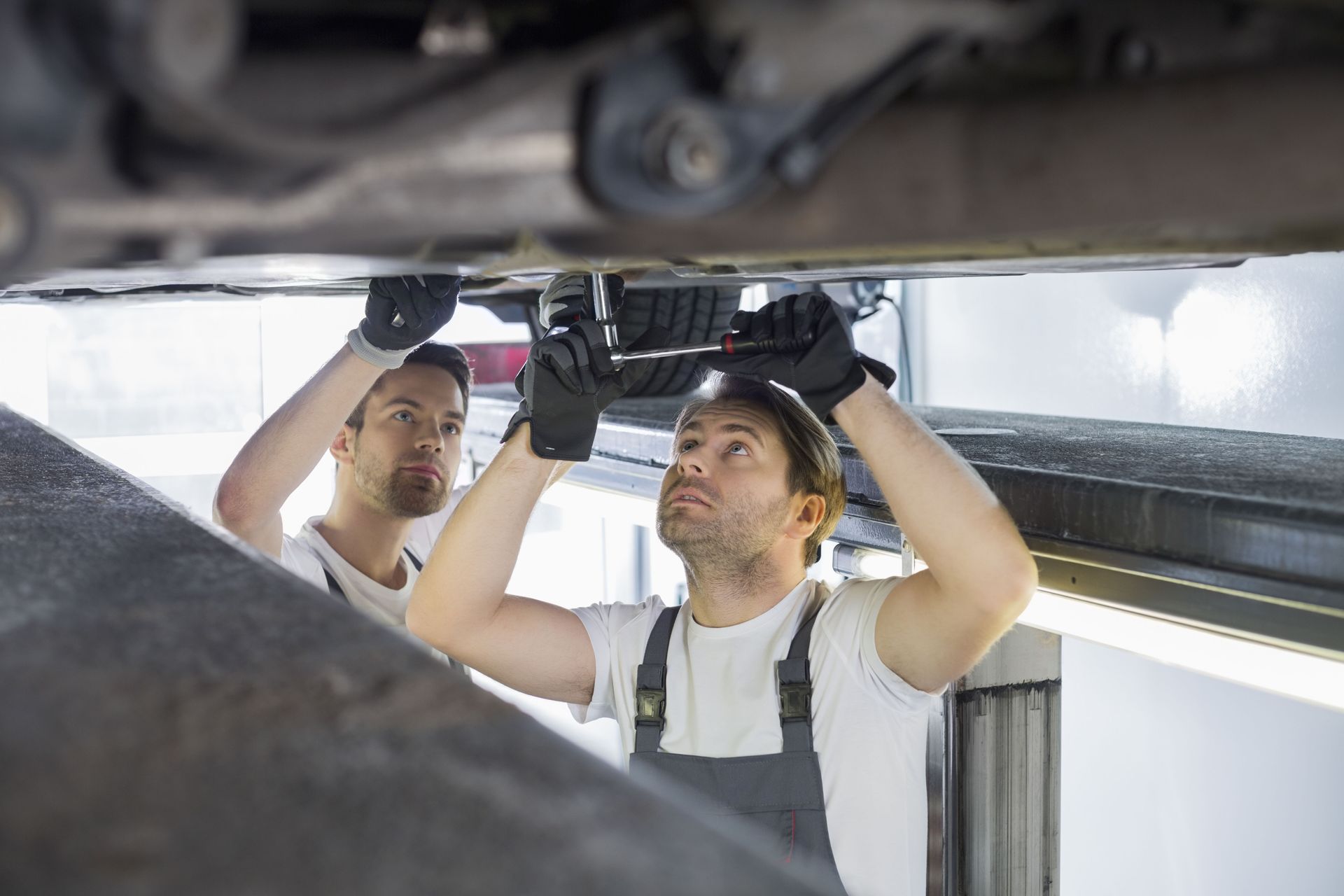 Two mechanics working under a car, one using a wrench. Bright garage setting.