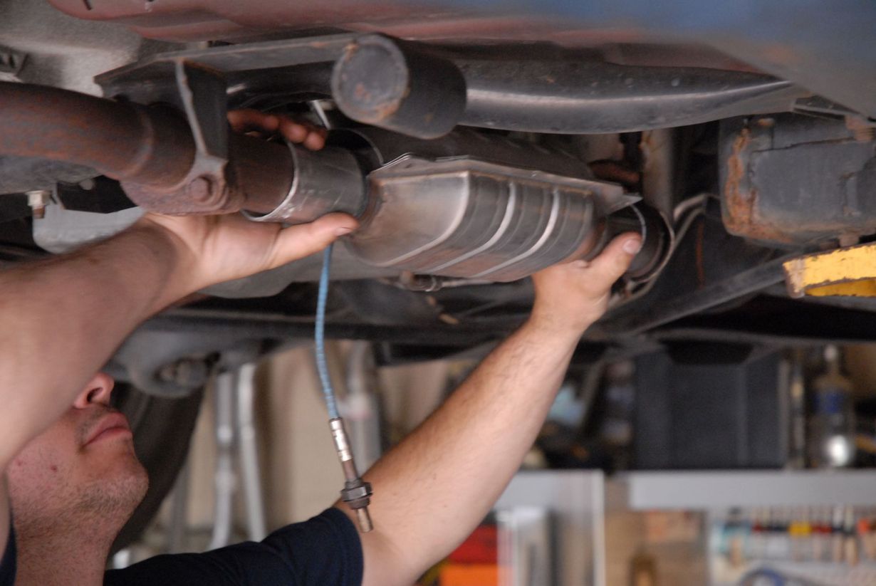 Man installs exhaust pipe under a car, connecting to a catalytic converter. Hands visible, shop setting.