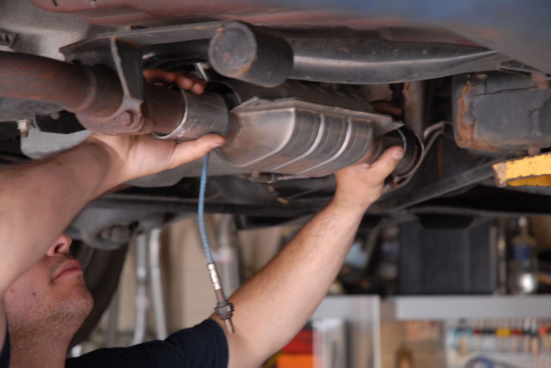 Man installs exhaust pipe under a car, connecting to a catalytic converter. Hands visible, shop setting.